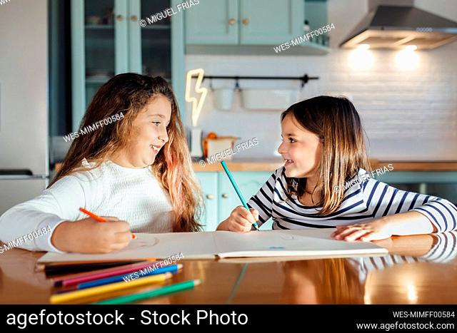 Smiling girls looking at each other while coloring with colored pencil on paper while sitting at dining table in kitchen