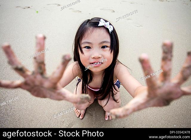 Young girl crouching on the beach and looking up at the camera to show her sandy hands; Hong Kong, China