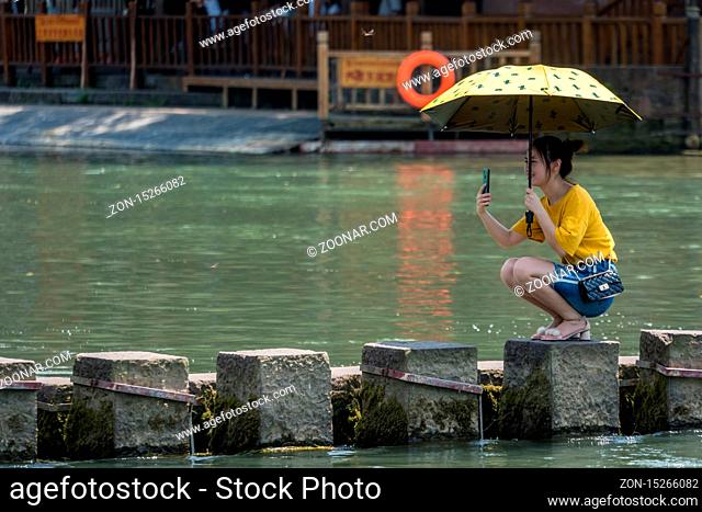 Feng Huang, China - August 2019 : Chinese girl with an umbrella squatting and taking selfie on her mobile on stepping stones on Tuojiang river