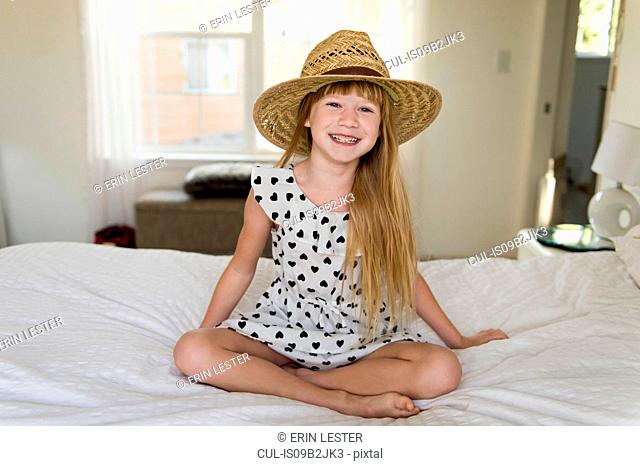 Young girl sitting on bed smiling, wearing straw hat