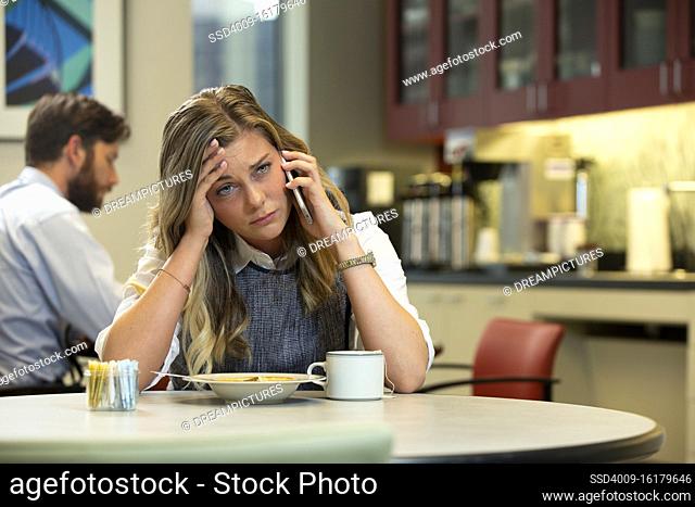 Young Caucasian woman with headache talking on cell phone while sitting in break area of office with lunch of soup and Hot Tea