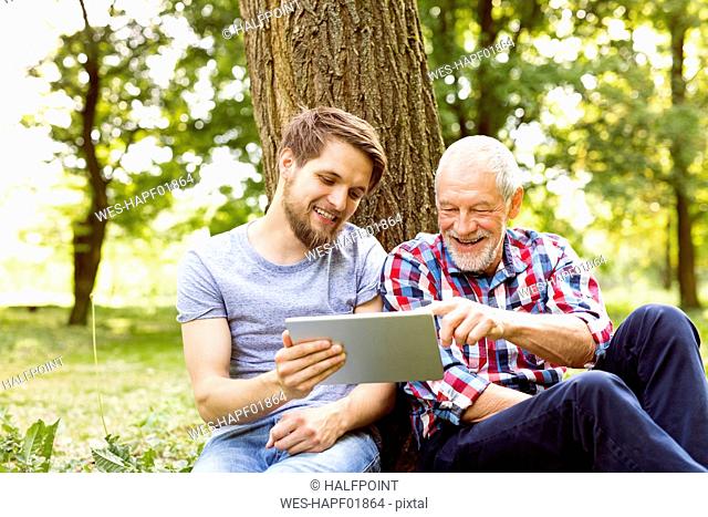 Happy senior father sitting besides his adult son in a park looking at tablet