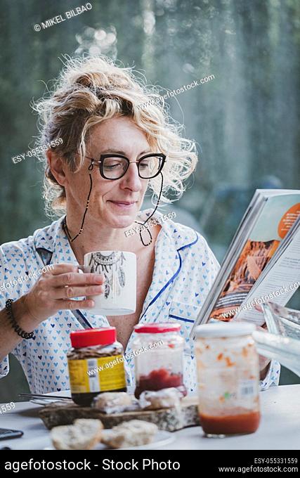 Blonde young mature woman with glasses in pyjamas at home in breakfast time, reading a magazine and having a cup of coffee