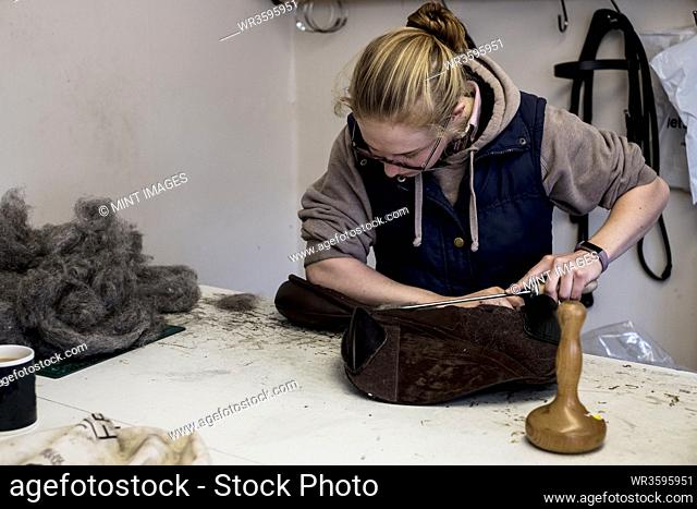 Female saddler standing in workshop, stuffing leather saddle with horse hair
