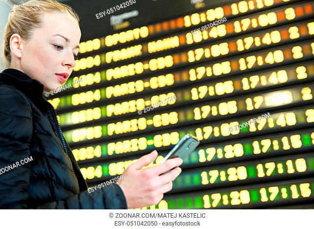 Young woman at international airport looking at the flight information board, holding smart phone in her hand, checking flight informations on phone application
