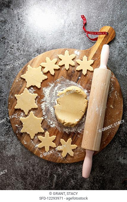 Cookie dough on a floured wooden chopping board surrounded with cutter out snowflake shapped biscuits next to a rolling pin on a grey slate background