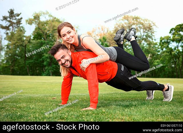 Sport man performing push-ups while woman sitting on his back. Happy couple training in green park or forest