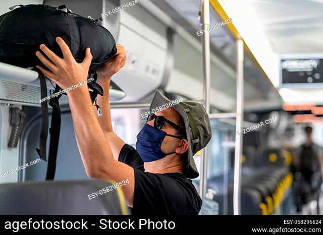 Middle aged male man wearing protective face mask, putting his backpack on above seat on a shelf in almost empty train carriage