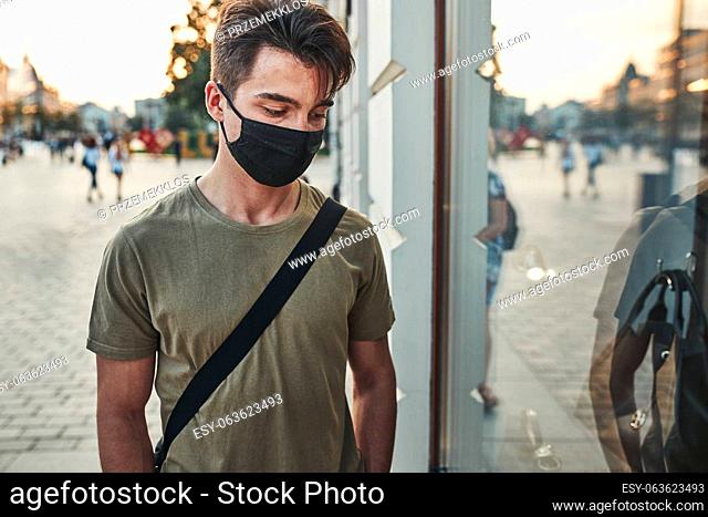 Young man walking along a store front in the city center in the evening wearing the face mask to avoid virus infection and to prevent the spread of disease in...