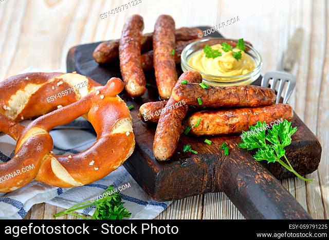 Acht gebratene Nürnberger Rostbratwürste mit Senf und Breze auf einem rustikalen Steakbrett serviert - Eight fried Bavarian sausages from Nuremberg with mustard...