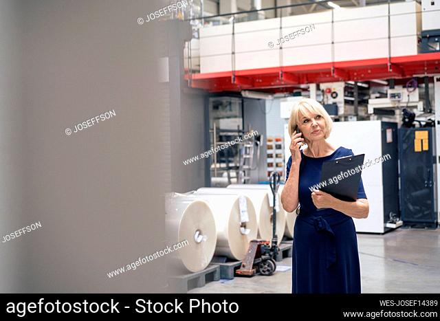 Senior businesswoman holding clipboard talking on mobile phone in factory