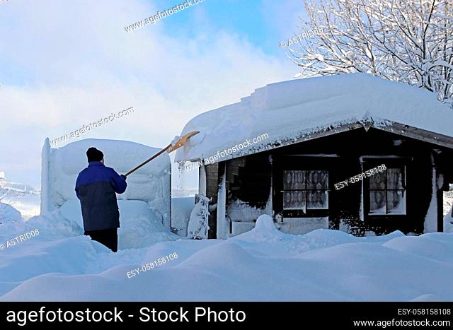 Man shovels high heavy snow from roof of wooden hut