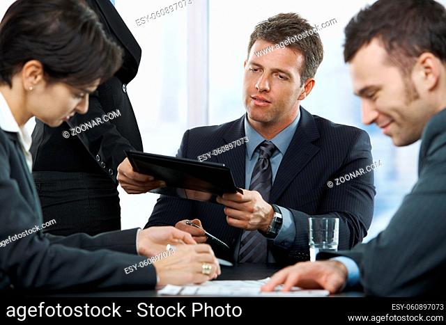 Executive getting report at meeting from assistant, other colleagues sitting looking down on documents