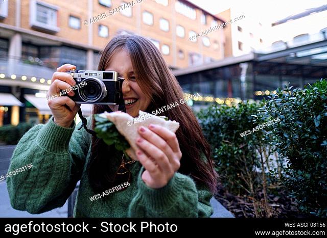 Happy woman holding sandwich photographing through camera in front of building