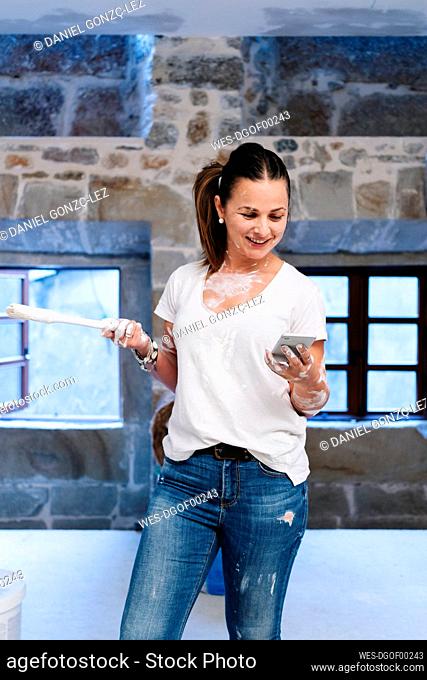 Woman using her cell phone while painting the walls of her new house