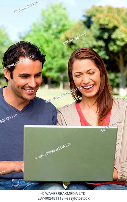 Two friends laughing as they watch something on a tablet while sitting together in a bright area surrounded by trees