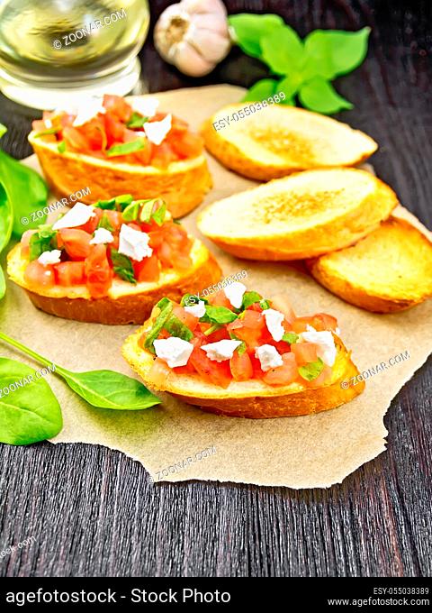 Bruschetta with tomato, spinach and soft cheese on paper, garlic, vegetable oil in a decanter and basil on wooden board background