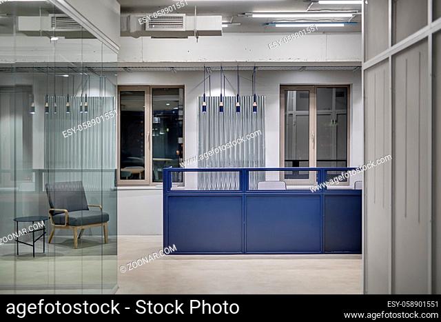 Blue metal reception rack with armchairs on the windows background in a loft style office with gray walls. On the left there is a gray armchair with a small...
