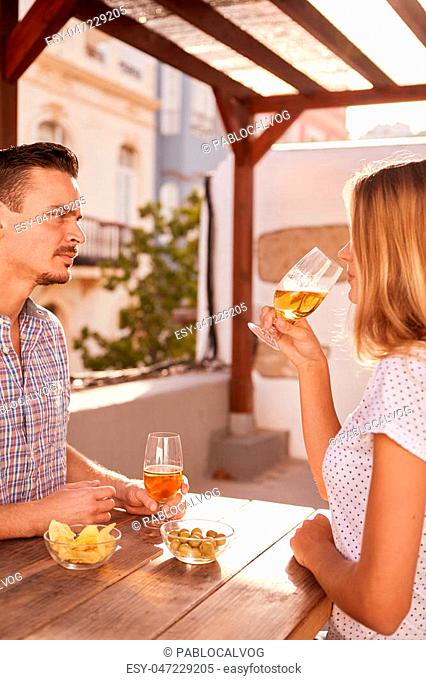 Good looking dark haired guy listening intently to blond girl while they are enjoying some drinks sitting at a table with snacks