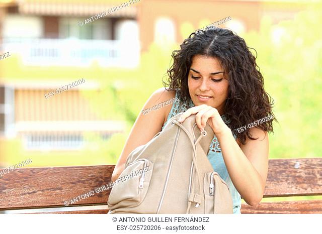 Woman searching something in her hand bag sitting in a bench of an urban park