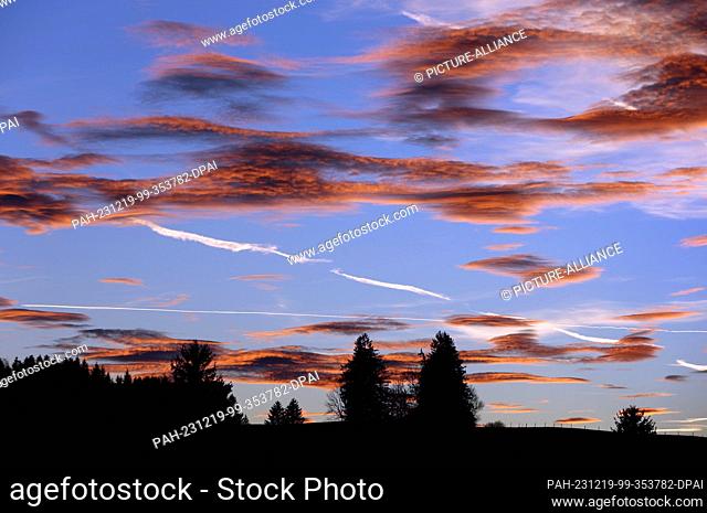 19 December 2023, Bavaria, Aitrang: The setting sun colors the clouds over the Allgäu landscape in different shades of red