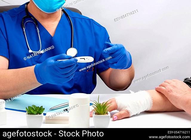 doctor sitting in blue uniform and latex gloves holds a white electronic thermometer, doctor’s office, reception of patients