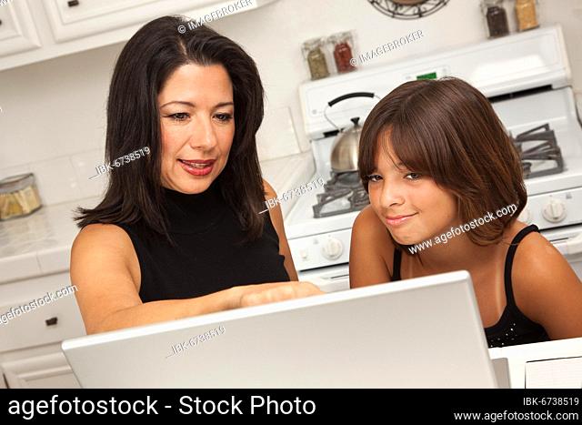 Attractive hispanic mother and mixed-race daughter in the kitchen using the laptop computer together