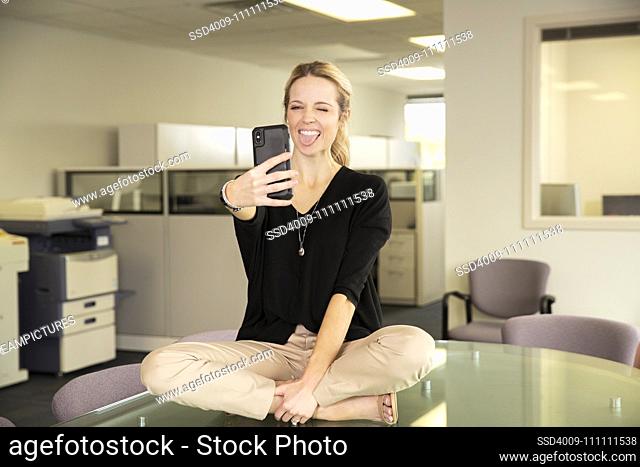 A young employee takes a selfie while sitting on top of a conference table in her office