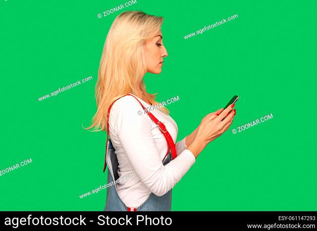Side view of adult pretty woman in stylish denim overalls holding smartphone and reading message, using mobile device for communication, browsing web