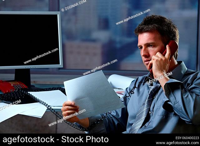 Mid-adult businessman talking on landline phone looking at business documents handheld