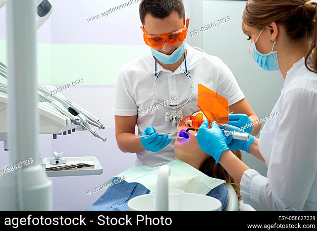 Female patient in blue shirt and patient bib on the patient chair in the dental cabinet. Next to her there is a male dentist and a female assistant