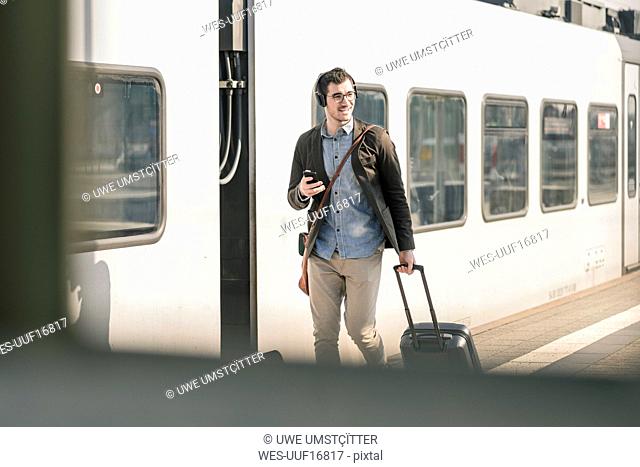 Smiling young man with headphones, cell phone and suitcase walking at station platform