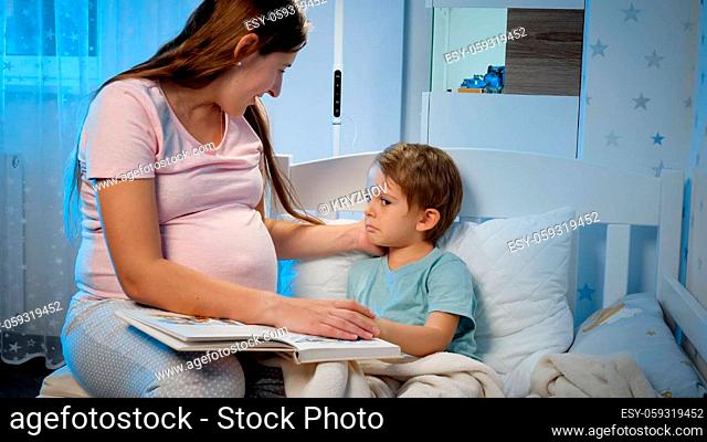 Upset and crying toddler boy lying in bed and hugging his pregnant mother sitting next to his bed at night