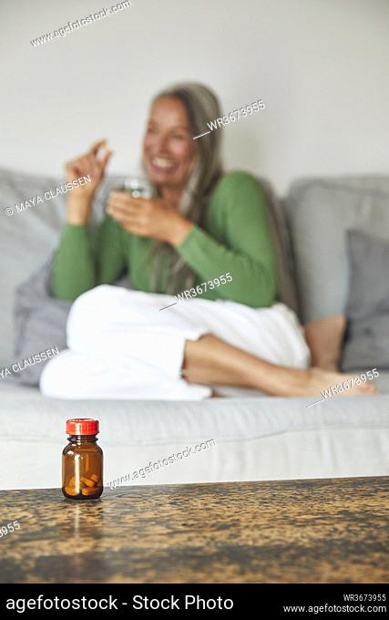 Smiling woman showing pill with drinking water on sofa at home
