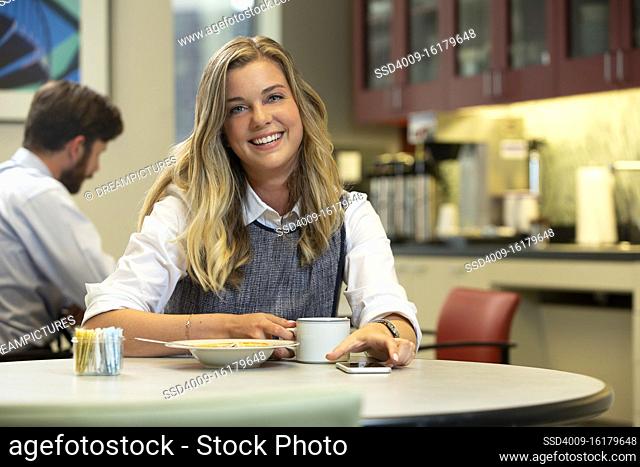Young Caucasian woman looking at camera with a smile, while sitting in break area of office with lunch of soup and Hot Tea
