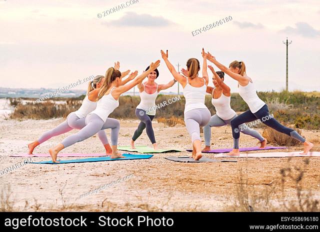 Group of slim attractive women wearing sportswear doing yoga standing on mats in circle outdoors on nature performing practising asana exercise Virabhadrasana...