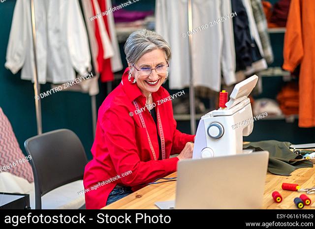 Video blog. Pretty adult woman in glasses smiling at laptop screen sitting at table with sewing machine material and threads
