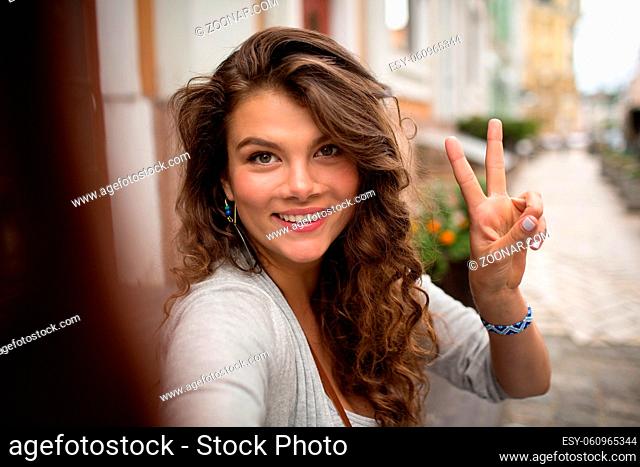 Young travel girl, having fun showing fingers on selfie photo. Woman walking in the street and making positive selfie shots