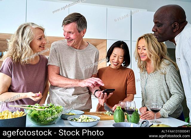 Diverse group of senior friends gathered at kitchen while using smart phone