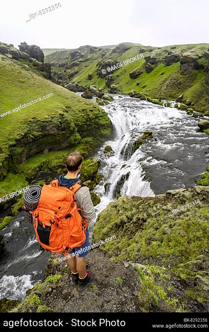 Hiker with large backpack in front of waterfall, rugged canyon overgrown with moss, river Skoga, landscape at Fimmvörðuháls hiking trail, South Iceland, Iceland