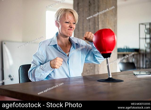Blond businesswoman punching boxing bag on table at home office