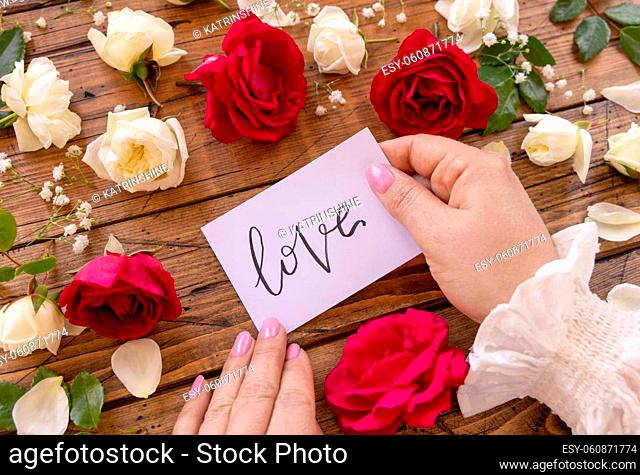 Hands with handwritten card LOVE surrounded by red and cream roses close up on a wooden table. Femminine romantic declaration of love near flowers