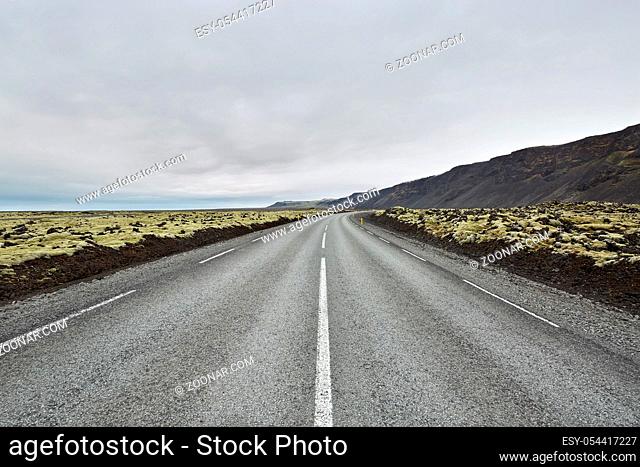 County roadway with orange roadside pillars between the green fields and mountains on the background of the cloudy sky in Iceland. Horizontal