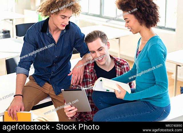 Three young students using both a paper book and a tablet PC for checking information during break in the classroom of a modern college