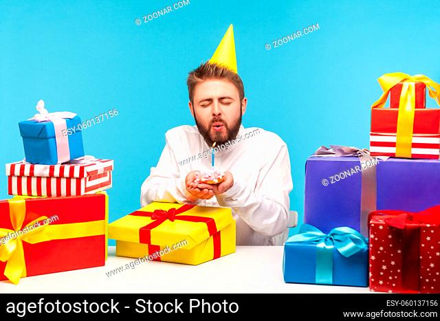 Funny bearded man in party cone making a wish closing eyes and blowing out candle on cake in his hands, celebrating birthday with many gifts at work