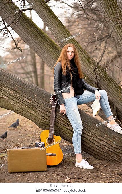 Red hair woman sitting on the tree exploring road map, guitar standing near, in the park. Travel concept