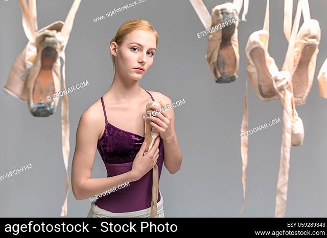 Cute ballerina in a violet top and a cream skirt stands on the gray background in the studio. In front of her there are hanging beige pointe shoes