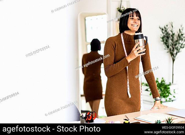 Happy young female stylist day dreaming while holding coffee cup in atelier