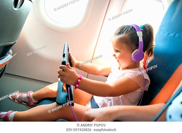 Adorable Little Girl With Headphone Over Her Head Using Digital Tablet During Traveling By Airplane