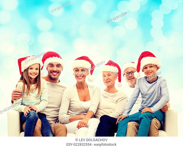 family, happiness, generation, holidays and people concept - happy family in santa helper hats sitting on couch over blue lights background